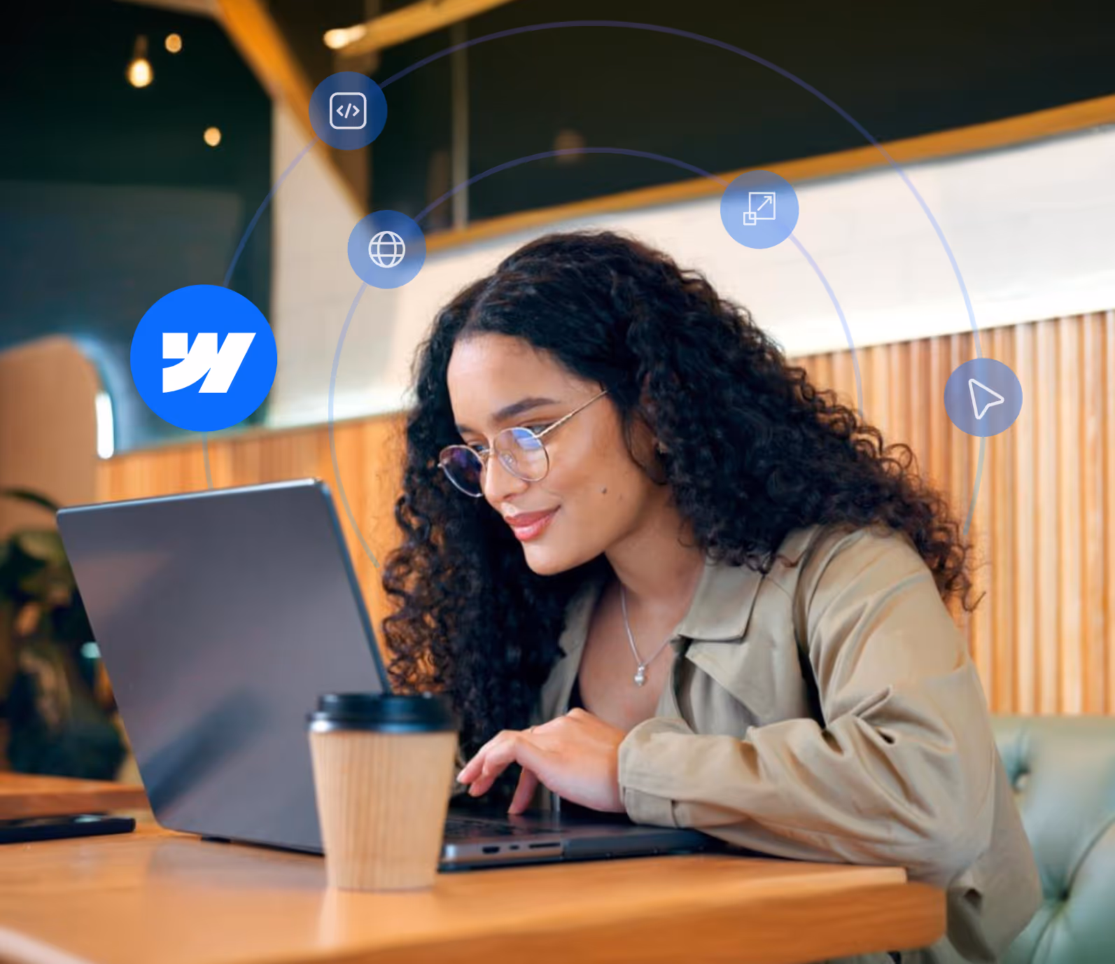 Woman with curly hair and glasses working on a laptop in a café with a coffee cup on the table and digital icons around her.