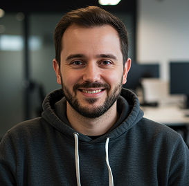 Smiling man with short dark hair and beard wearing a dark hoodie in an office setting.