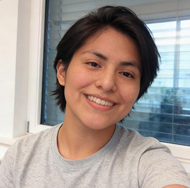 Smiling person with short dark hair wearing a light gray shirt sitting indoors near a window with blinds.