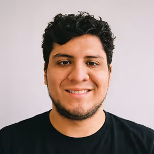 Smiling man with curly dark hair and a trimmed beard wearing a black shirt against a plain light background.