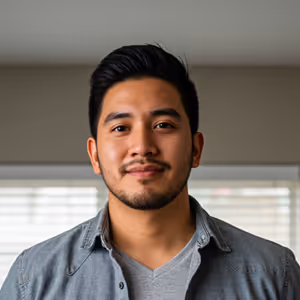 Portrait of a young man with dark hair and beard wearing a gray shirt and jacket, standing indoors with a window in the background.