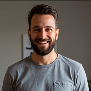 Smiling bearded man with dark hair wearing a gray t-shirt against a blurred indoor background.
