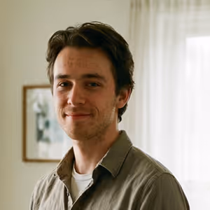 Smiling young man with dark hair wearing a brown shirt, standing indoors with a framed picture and sheer curtains in the background.