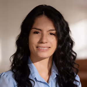 Smiling woman with long curly dark hair wearing a light blue shirt, in soft indoor lighting.