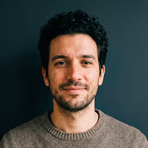 Smiling man with curly dark hair and beard wearing a brown sweater against a dark background.