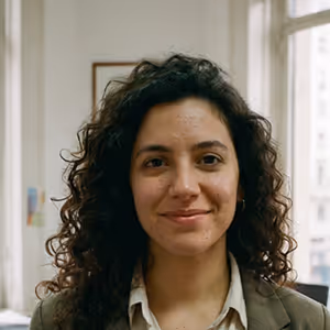 Smiling woman with curly dark hair wearing a blazer and shirt, standing indoors with windows behind her.