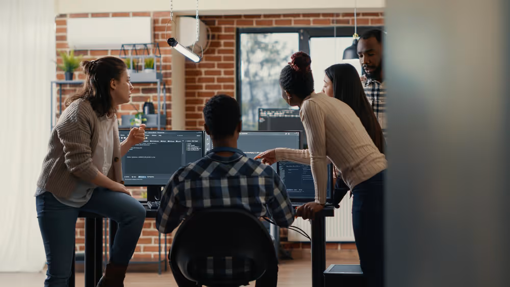 Five diverse coworkers collaborating around two computer monitors in a modern office with brick walls.