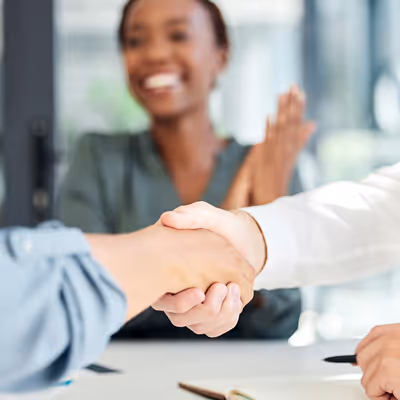Two people shaking hands in the foreground with a smiling person clapping in the blurred background.