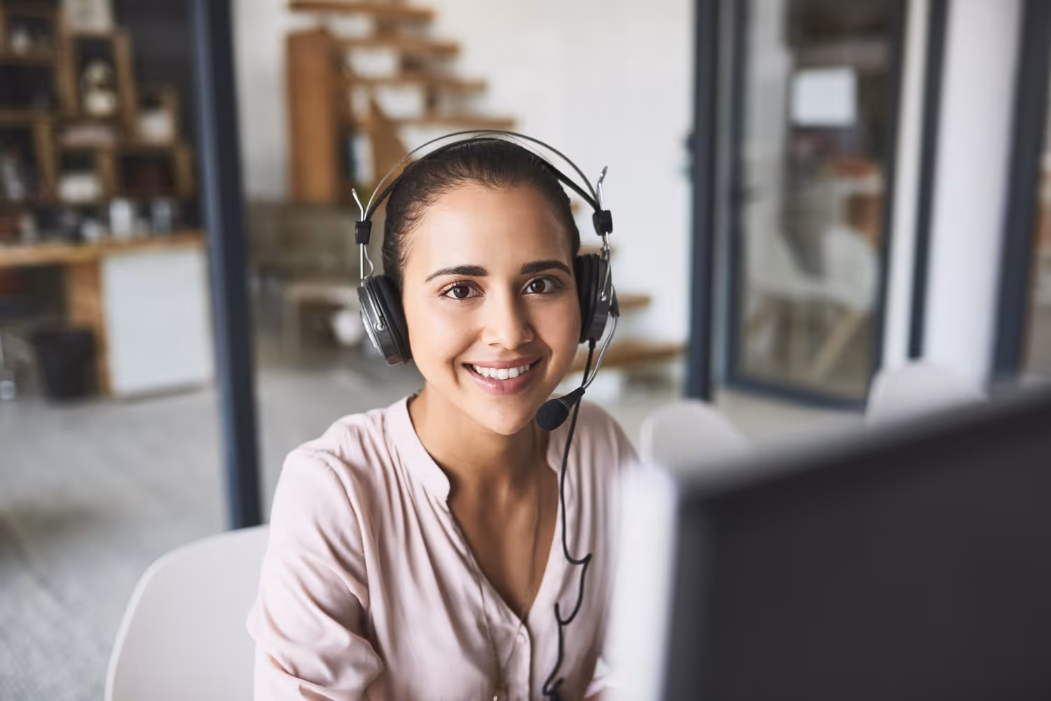 Smiling woman wearing headset with microphone sitting at a desk in a modern office.
