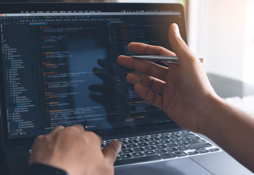 A person pointing at code on a laptop screen with a pen while typing with the other hand.