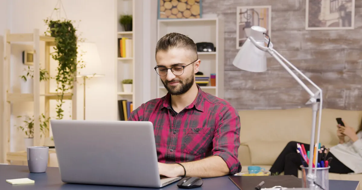 Man with glasses and a red plaid shirt works on a laptop at a desk; another person sits on a couch in the background using a smartphone.