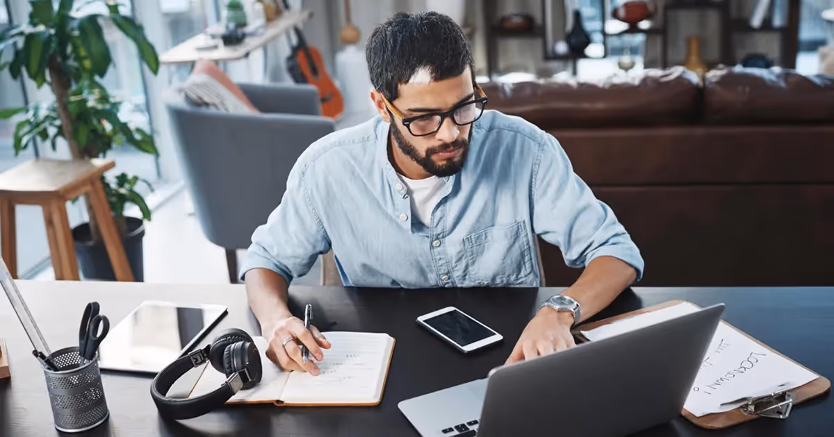 Man wearing glasses works at a laptop on a desk, taking notes in a notebook. Headphones, a tablet, and a smartphone are also on the desk.