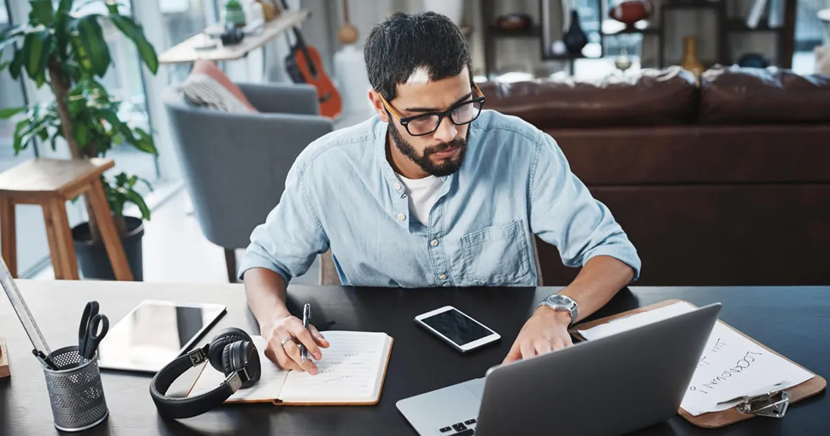 Man wearing glasses works at a laptop on a desk, taking notes in a notebook. Headphones, a tablet, and a smartphone are also on the desk.