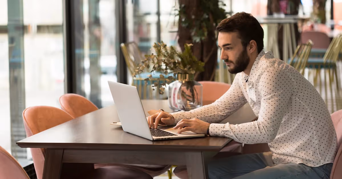 A man seated at a table, focused on his laptop, with a neutral background.
