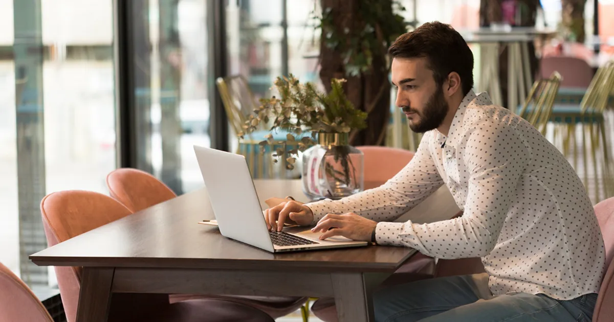 A man seated at a table, focused on his laptop, with a neutral background.