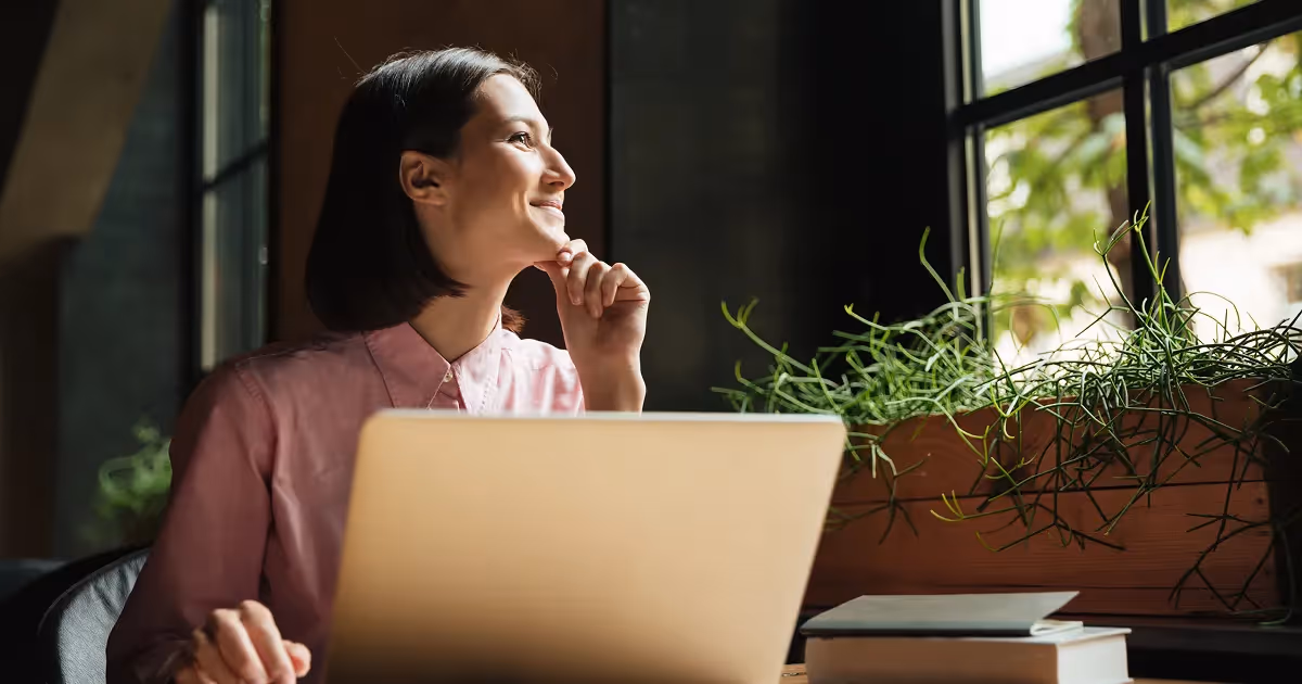 A woman sitting at a table, focused on her laptop, with a cozy indoor setting in the background.