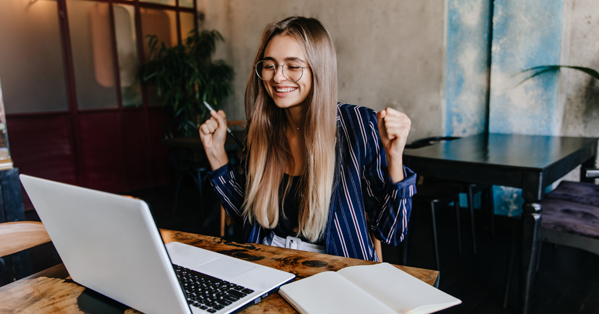 Young woman with long hair and glasses, excitedly looking at a laptop in a cozy cafe. She raises her fists in celebration, an open notebook nearby.