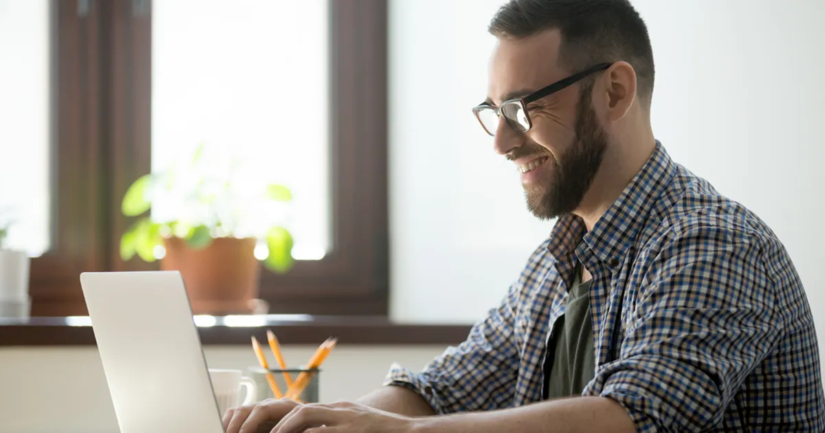 A bearded man with glasses smiles while typing on a laptop in a bright room. A potted plant by the window adds a touch of greenery.