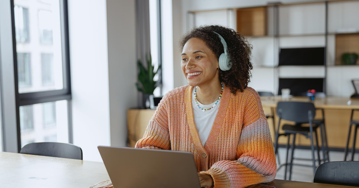 A smiling woman wearing headphones and a colorful sweater sits at a table with a laptop in a bright, modern workspace. The atmosphere is relaxed and cheerful.