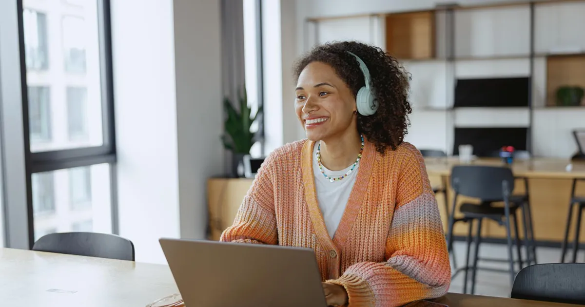 A smiling woman wearing headphones and a colorful sweater sits at a table with a laptop in a bright, modern workspace. The atmosphere is relaxed and cheerful.
