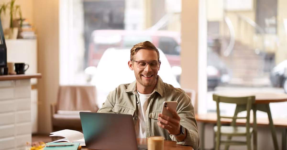 A man with glasses, smiling and holding a smartphone, sits at a cafe table with a laptop and coffee. The setting is cozy and inviting.