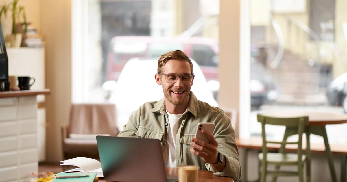 A man with glasses, smiling and holding a smartphone, sits at a cafe table with a laptop and coffee. The setting is cozy and inviting.