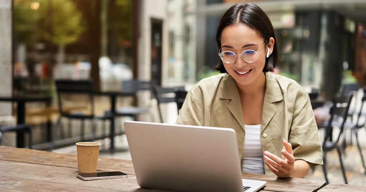 A woman with glasses sits in a café, engaged in a video call on her laptop. She is smiling, conveying a friendly and relaxed atmosphere. A coffee cup and phone are on the table.