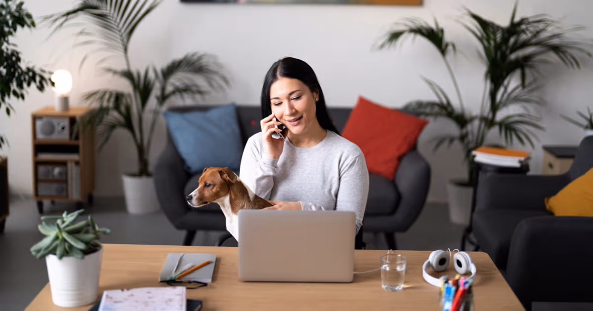 A woman sits at a desk with a laptop, while a dog rests beside her, creating a cozy work environment.