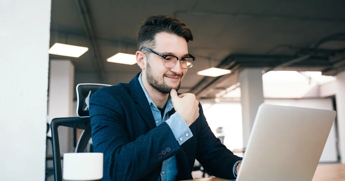 A man wearing glasses sits at a desk, focused on his laptop.