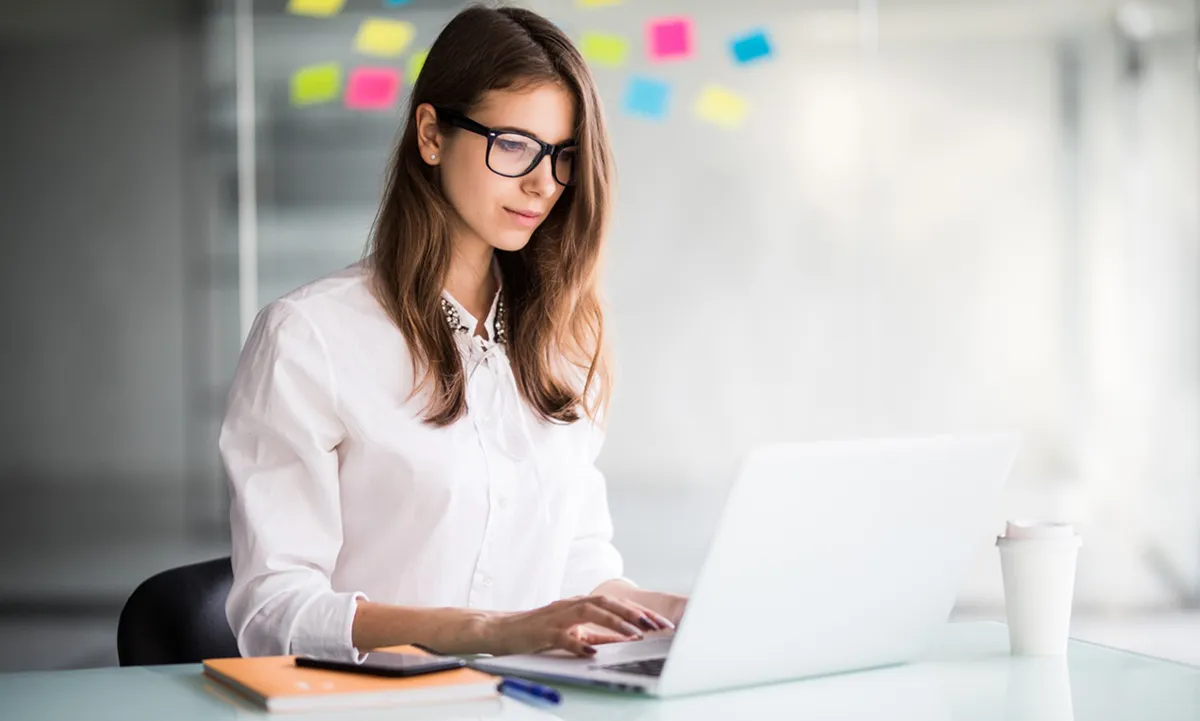 A woman wearing glasses is focused on her laptop, engaged in work at a desk.