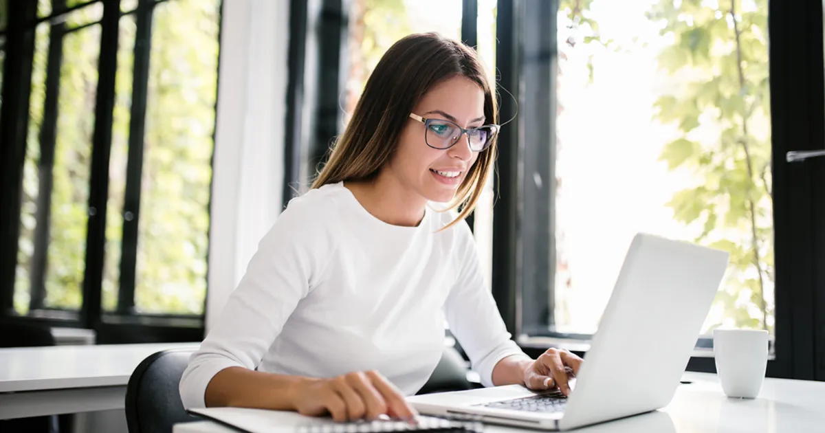 A woman wearing glasses is focused on her laptop, engaged in work at a desk.