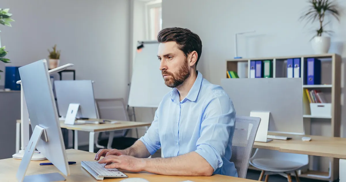 A man sits at a desk, focused on his computer screen, with papers and a pen nearby.