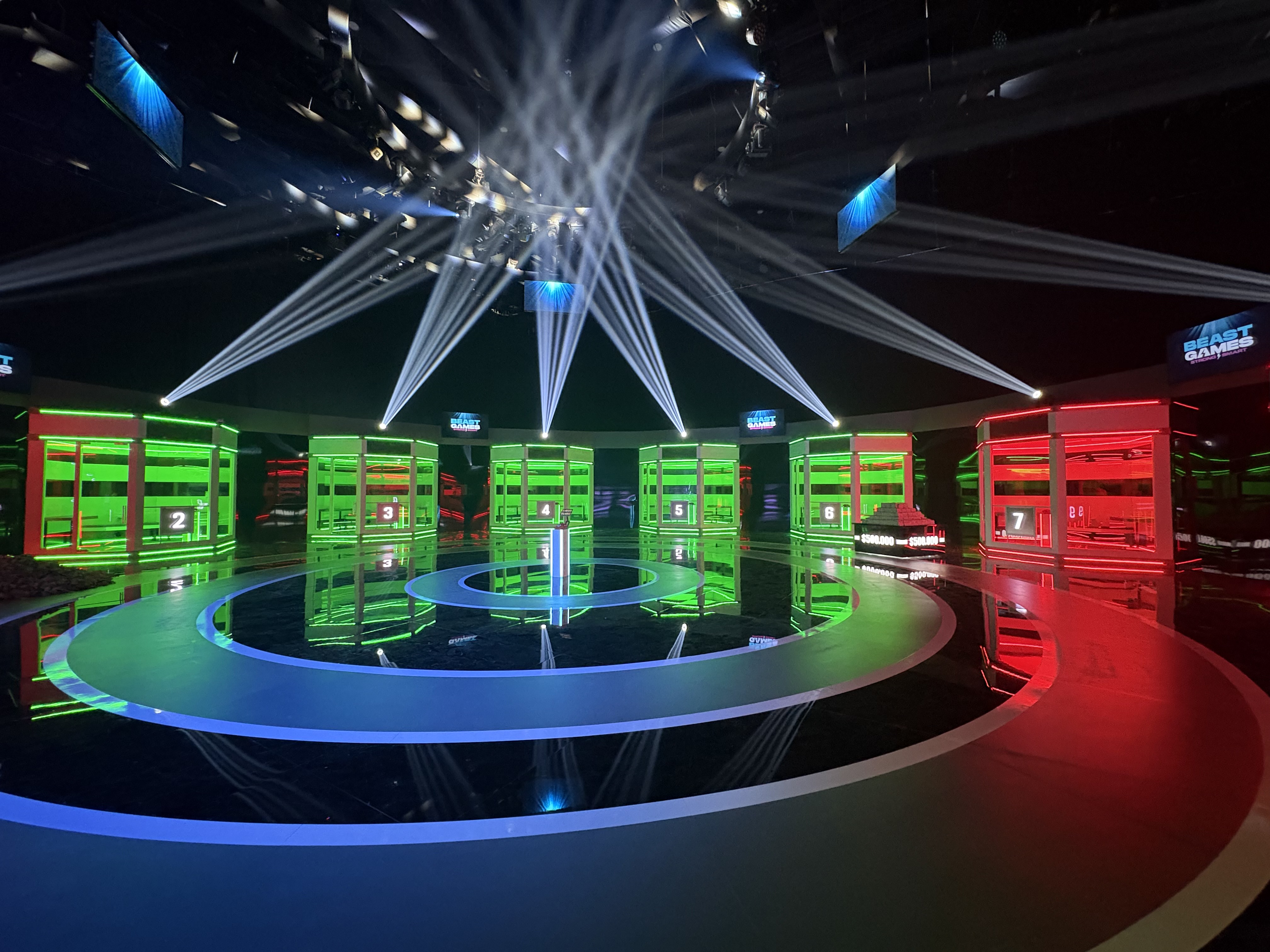 Inside a soundstage, an illuminated platform is centered. Four-green lighted and one-red lighted room sit behind the platform.
