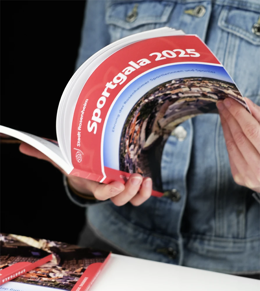 Eine Person in einer Jeansjacke hält eine aufgeschlagene Broschüre mit dem Titel „Sportgala 2025“ und rotem Einband in der Hand. Der dunkle Hintergrund betont die Heftfarbe.