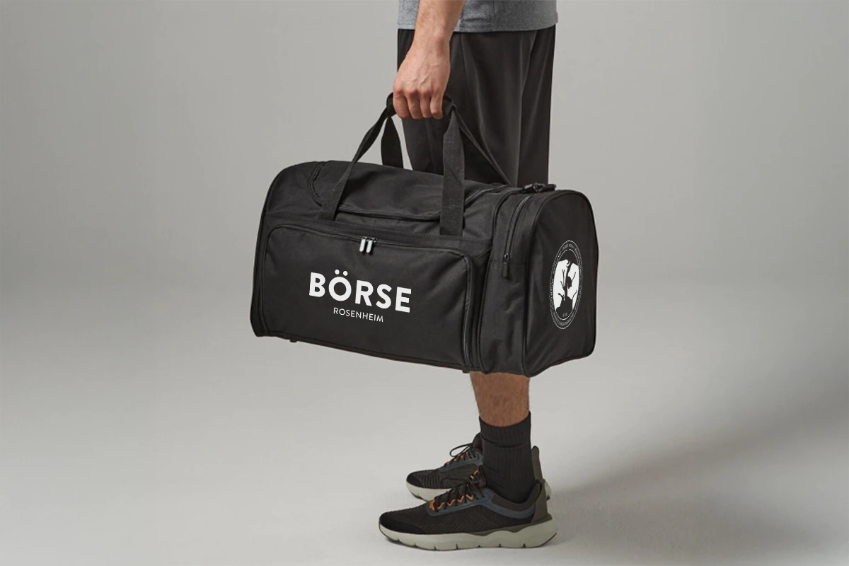 Eine Person hält eine schwarze Reisetasche mit der Aufschrift „BÖRSE ROSENHEIM“ vor einem schlichten grauen Hintergrund. Sie trägt Sportshorts und Turnschuhe. Der Ton ist lässig.
