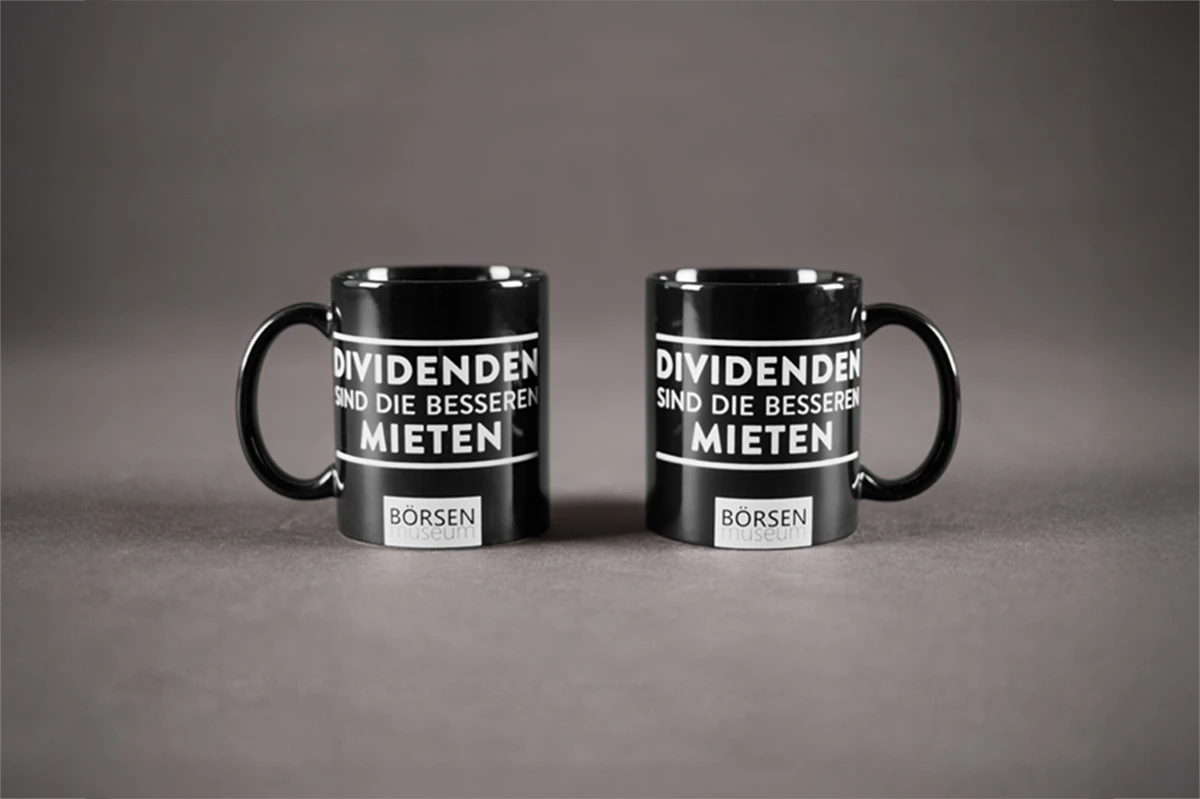 Zwei schwarze Tassen mit weißem Text „Dividenden sind die besseren Mieten“ auf grauem Hintergrund.