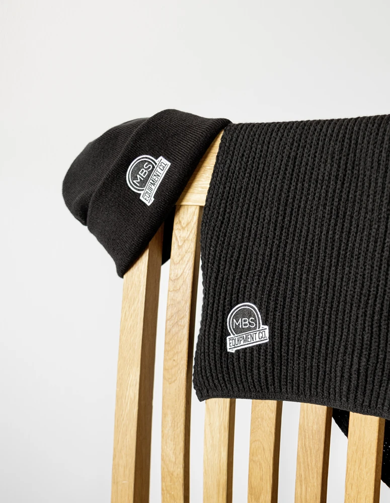 Schwarze Mütze und Schal mit dem Logo „MBS Equipment Co.“ über einem Holzstuhl vor hellem Hintergrund, was Schlichtheit und Wärme vermittelt.