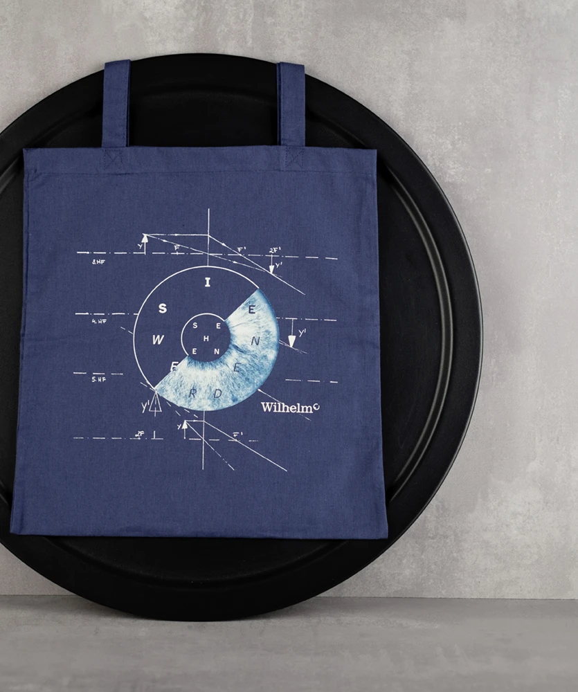 Blaue Einkaufstasche mit abstraktem weiß-blauem Kreismuster und geometrischen Linien vor einer grauen Wand. Der Text lautet „Wilhelm“. Modern und stilvoll.