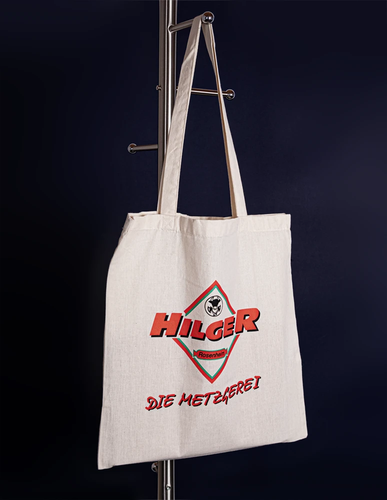 Vor dunklem Hintergrund hängt eine beigefarbene Einkaufstasche an einem Ständer. Auf der Tasche ist in kräftigen roten Buchstaben der Text „HILGER DIE METZGEREI“ und ein Logo zu sehen.