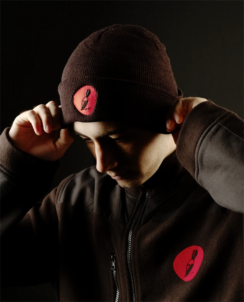 Junger Mann trägt eine schwarze Mütze und schwarze Jacke, die jeweils mit einem roten Logo mit Pinsel gebrandet sind. Dunkle, misteriöse Stimmung