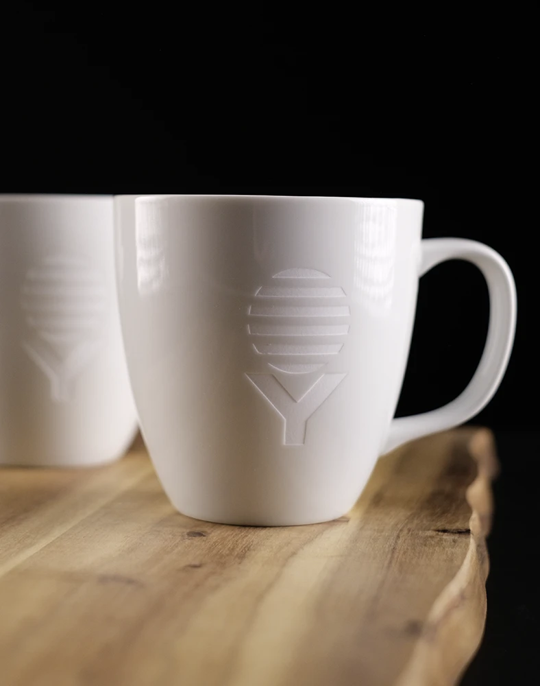 Weiße Tasse mit erhabenem Logo in Form von horizontalen Linien und einem 'Y' auf der Vorderseite, auf einem Holzbrett platziert.