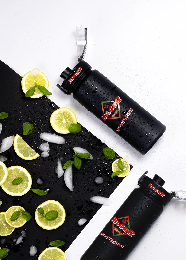 Zwei schwarze Trinkflaschen mit dem Aufdruck 'HILGER' liegen auf einer dunklen Oberfläche, umgeben von Zitronenscheiben, Minzblättern und Eiswürfeln.