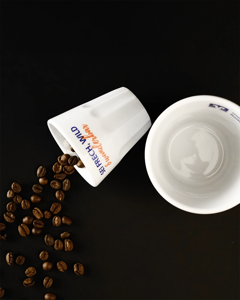 Zwei weiße Tassen, eine umgekippt mit Kaffeebohnen, die herausfallen. Auf einer Tasse steht 'SERIE: ACHT MIND'. Die andere Tasse ist leer.