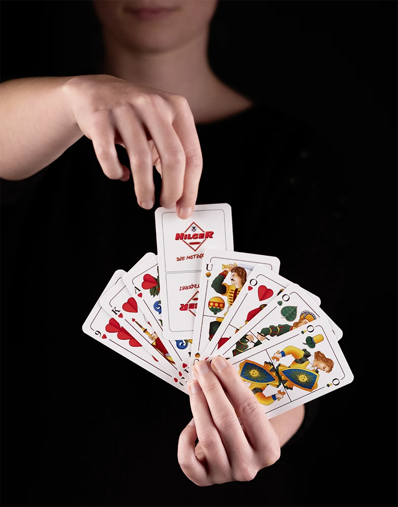 Eine Person hält ein Fächer aus Spielkarten mit Herzsymbolen und Figuren. Eine Karte zeigt das Logo 'Hilger'. Die Person hat eine schwarze Kleidung und eine Hand hebt die Karten.