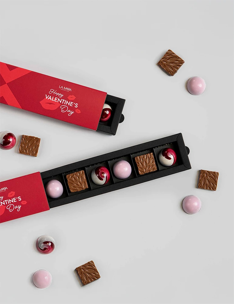 Schachtel mit Pralinen von La Mara, rot mit dem Text 'Happy VALENTINE'S Day', enthält sechs Pralinen in verschiedenen Farben und Formen, umgeben von weiteren Pralinen.