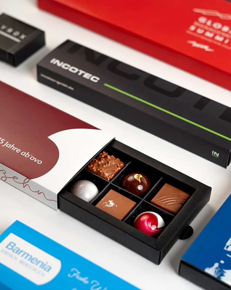 Schwarze Schachtel mit sechs Pralinen in verschiedenen Formen und Farben, umgeben von weiteren Verpackungen in verschiedenen Farben.