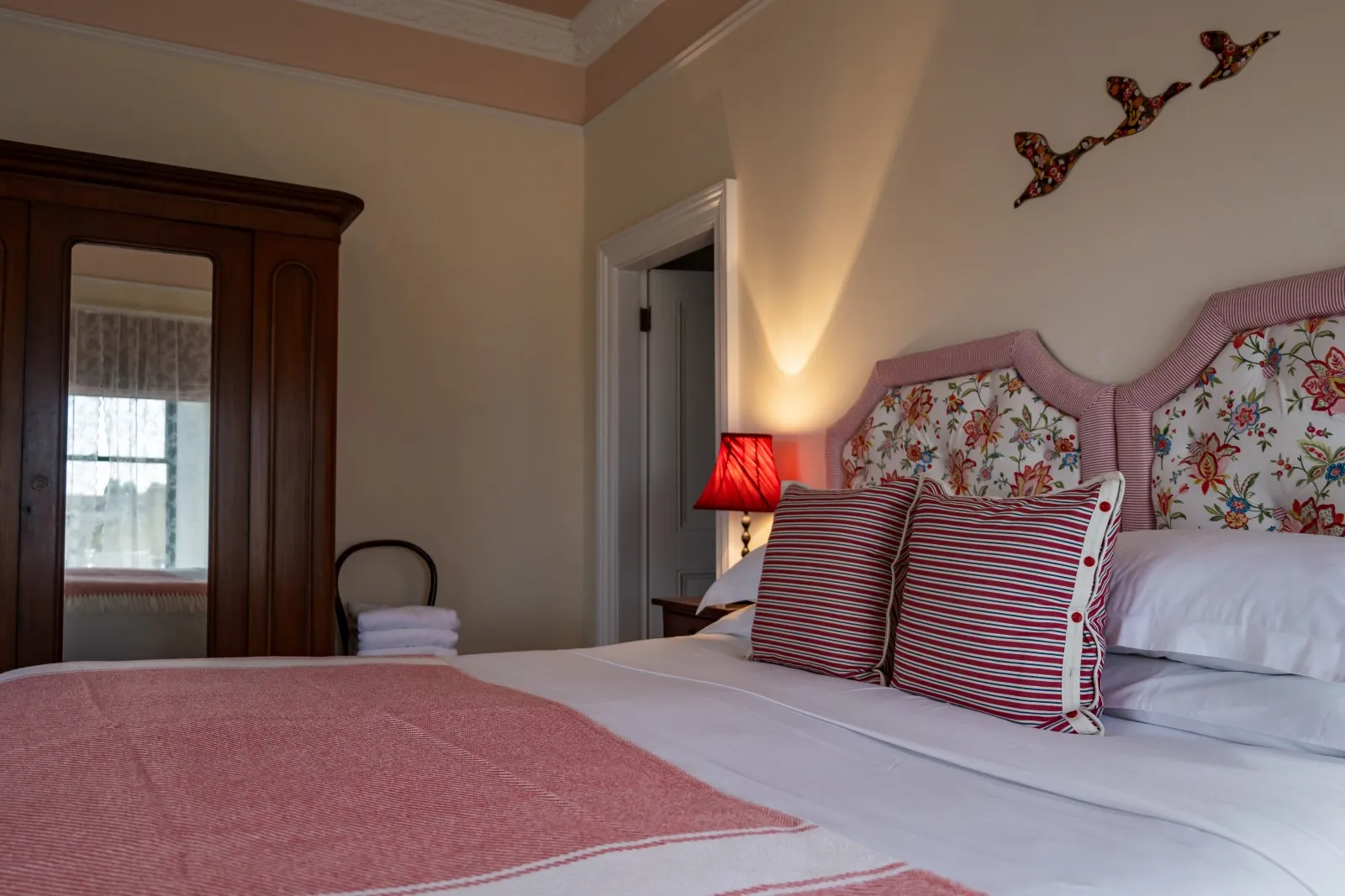 Bed with floral and striped pillows, red throw, wooden wardrobe, and red lamp in cozy bedroom.