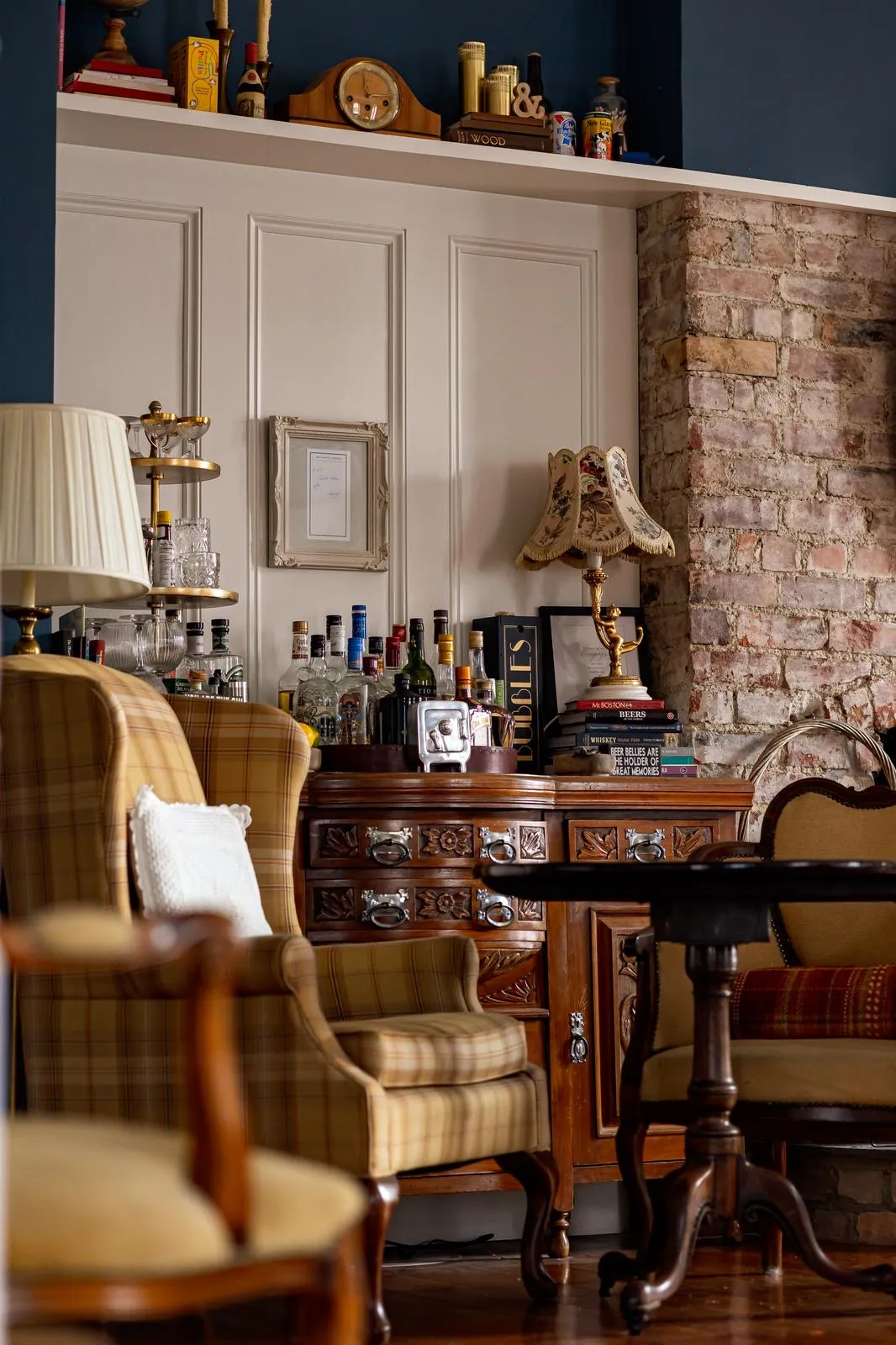 Cozy vintage sitting area with plaid armchair, wooden table, brick wall, and cabinet holding bottles and a floral lamp.