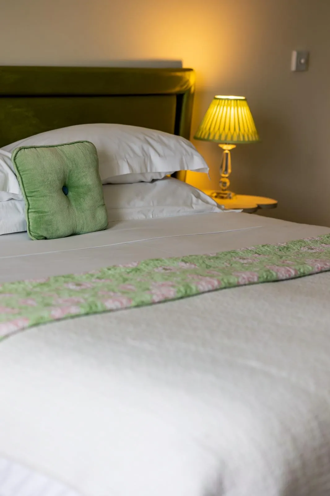 Neatly made bed with white sheets, green pillow, patterned green and pink runner, and a lit bedside lamp.