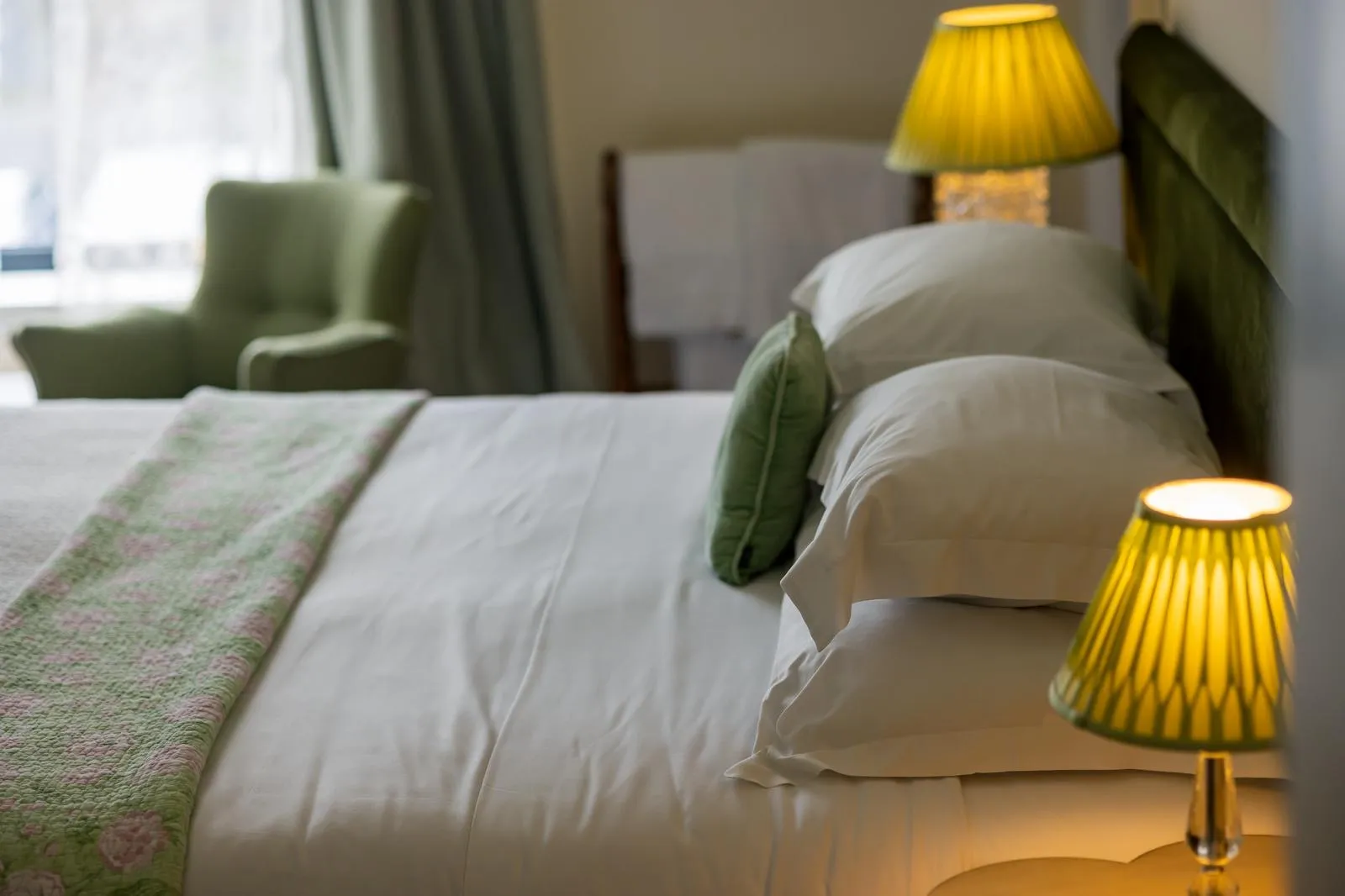 Close-up of a neatly made bed with white sheets, green pillows, and a green-patterned blanket, lit by yellow lamps.